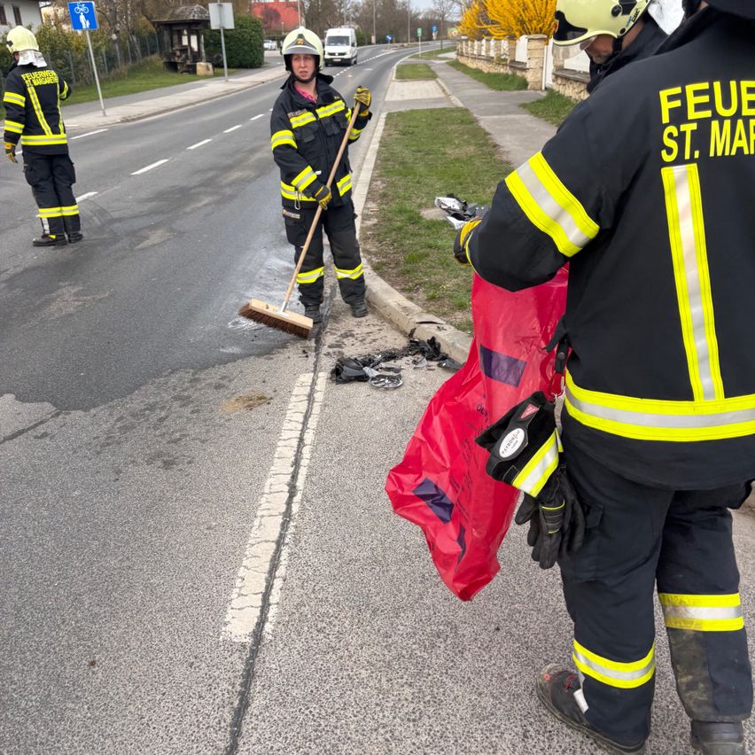 Drei Feuerwehrleute räumen Trümmer auf der Straße auf. Einer hält einen Besen, der andere einen roten Beutel. Sie tragen reflektierende Westen und Helme.