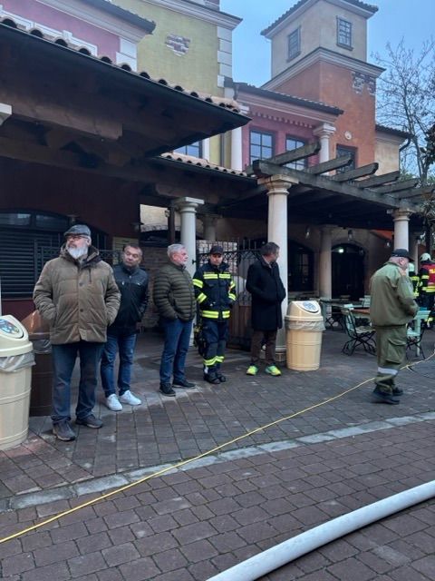 Eine Gruppe von Menschen in Jacken steht vor einem Gebäude mit Feuerwehrleuten, einer hält eine Feuerwehrhose. In der Nähe befinden sich Mülleimer und Stühle.