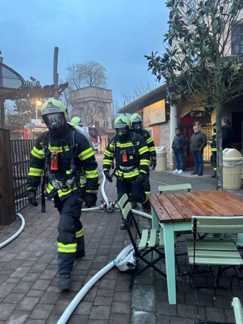 Feuerwehrleute in voller Ausrüstung, einschließlich Helmen und Masken, gehen an einem Gebäude mit Stühlen und Tischen vorbei. Ein Feuerwehrschlauch liegt auf dem Boden, und Menschen stehen herum.