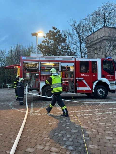 Ein Feuerwehrmann geht mit einem Schlauch in der Hand, und zwei andere stehen bei einem Feuerwehrwagen.