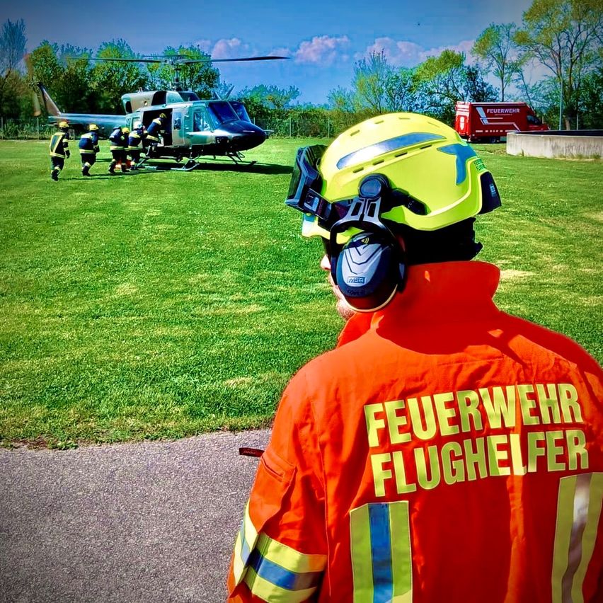 Ein Feuerwehrmann in einer orangefarbenen Uniform mit den Worten 'Feuerwehr' und 'Flughelfer' steht vor einem Hubschrauber, während andere Feuerwehrleute ihn besteigen. Der Hubschrauber ist auf einem Rasenfeld geparkt, in der Nähe eines roten Lkw.