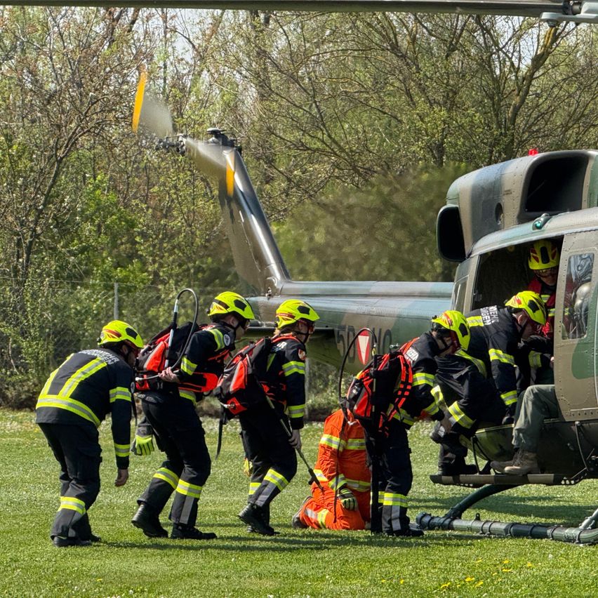 Feuerwehrleute in gelben und schwarzen Uniformen bereiten sich darauf vor, einen Militärhubschrauber auf einem Rasenfeld zu besteigen. Bäume und ein klarer Himmel sind im Hintergrund sichtbar.