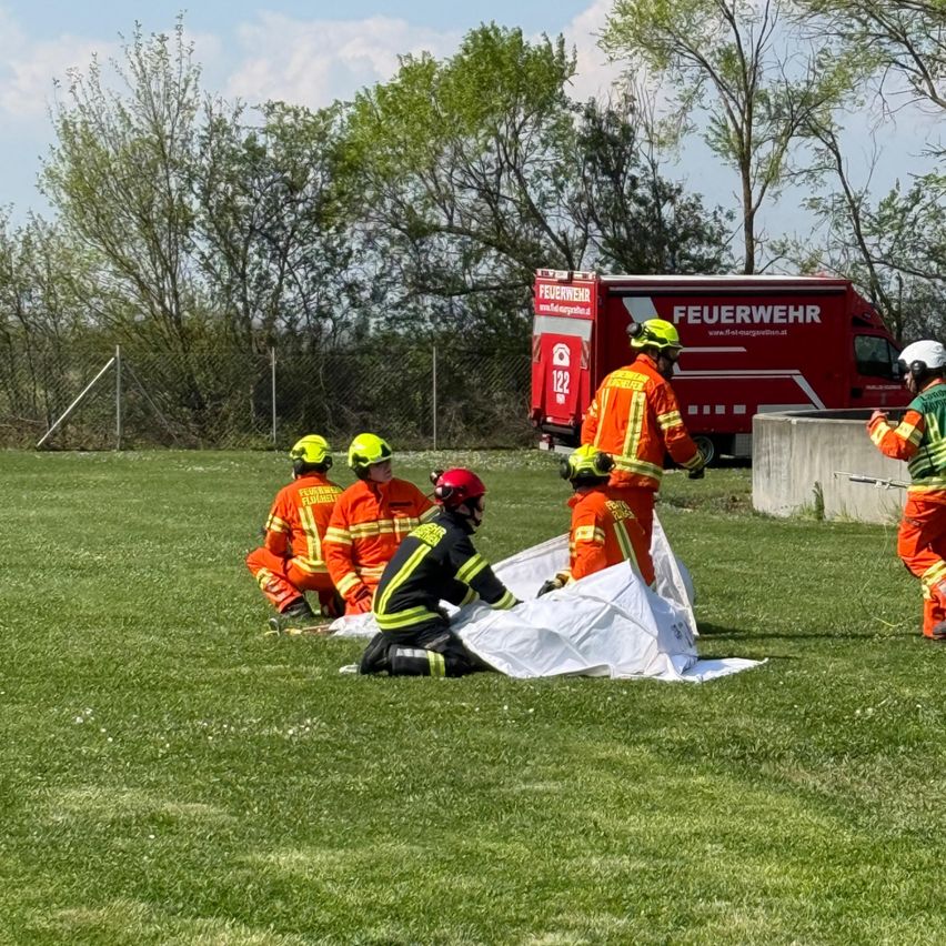 Mehrere Feuerwehrleute trainieren auf einem grünen Feld. Ein Feuerwehrmann hält eine Plane, während andere zusehen. Ein roter Feuerwehrwagen steht hinter ihnen.