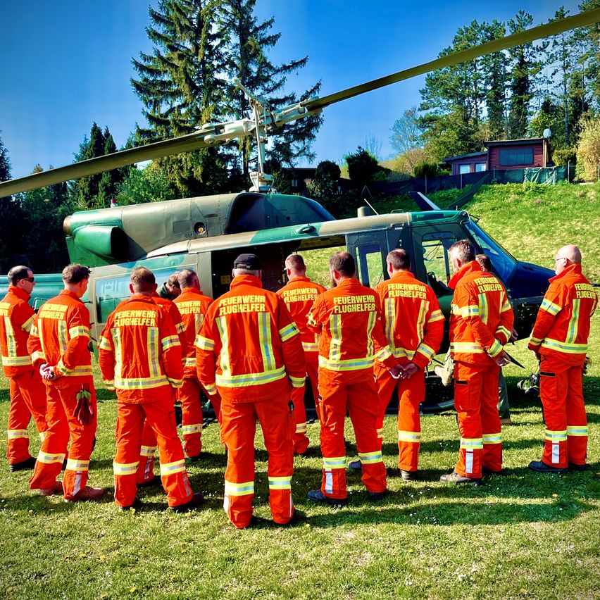 Eine Gruppe Feuerwehrleute in orangefarbenen Uniformen steht vor einem Hubschrauber auf einem Rasen. Der Hubschrauber hat die Aufschrift 'Feuerwehr' an der Seite.