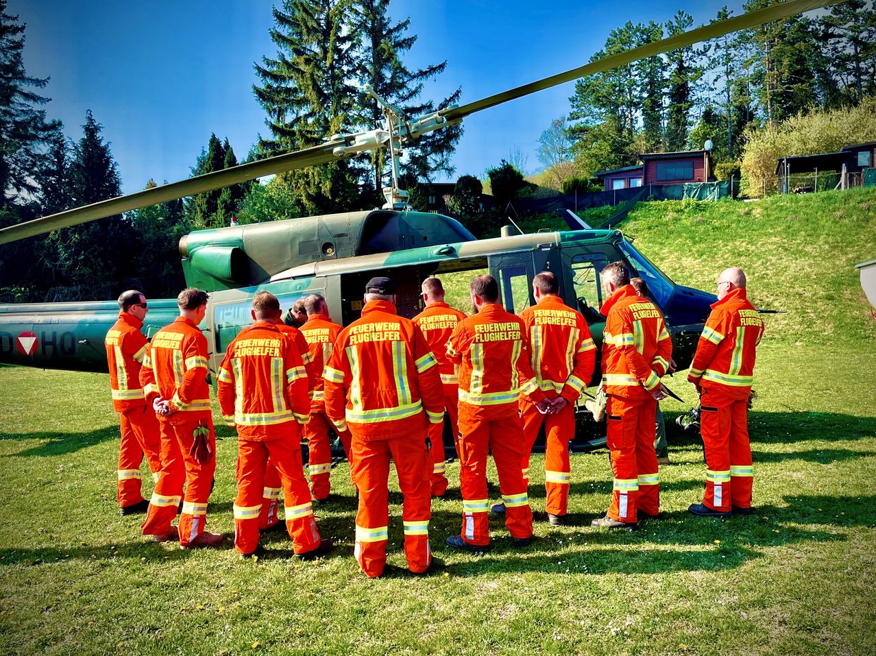Eine Gruppe Feuerwehrleute in orangefarbenen Uniformen steht vor einem Hubschrauber auf einem Rasen. Der Hubschrauber hat die Aufschrift 'Feuerwehr' an der Seite.