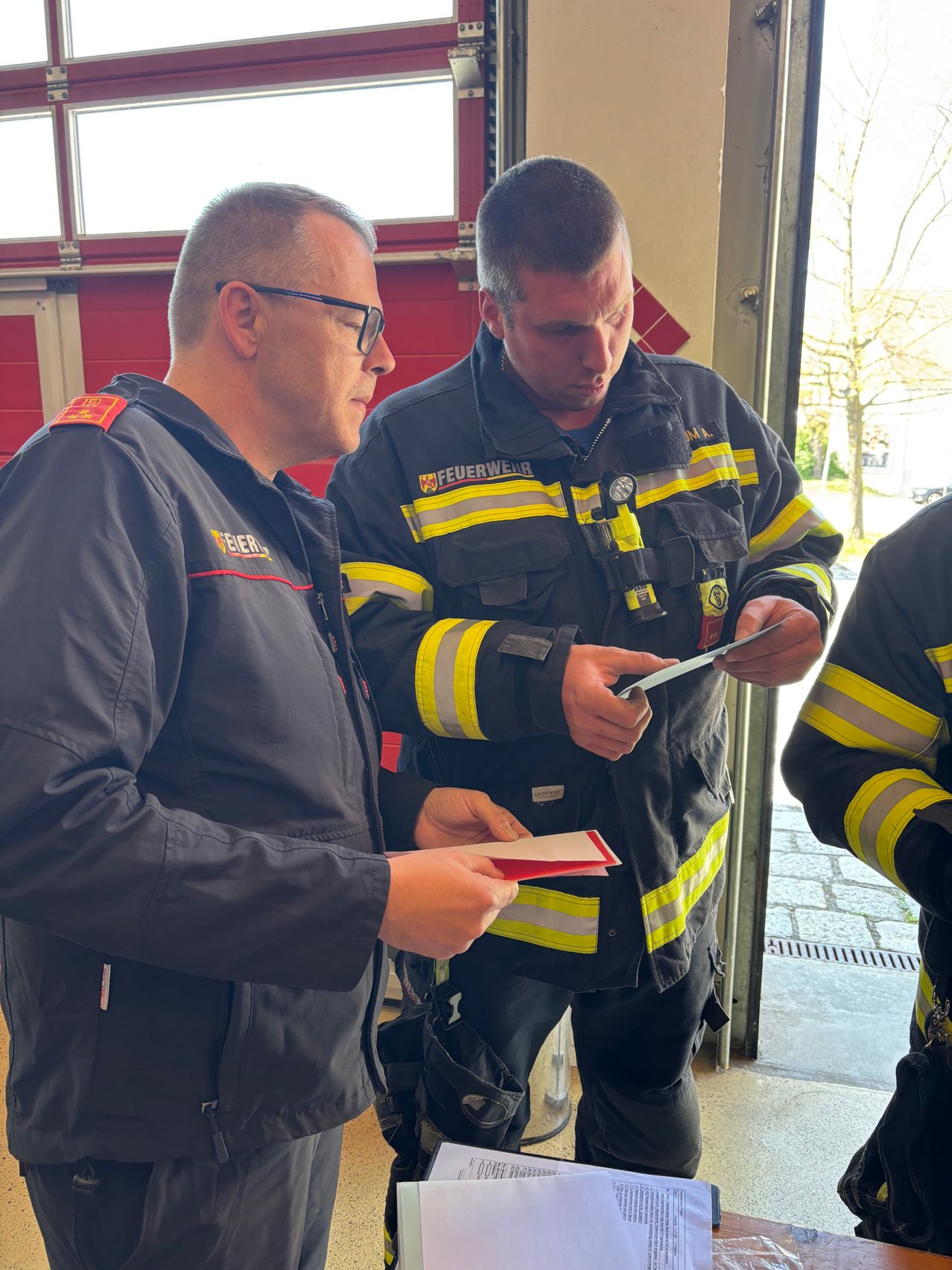 Zwei Feuerwehrleute in Uniform, einer hält ein Stück Papier in der Hand, besprechen etwas, während sie vor einer Feuerwache stehen.