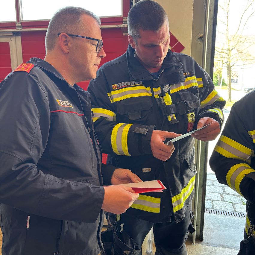 Zwei Feuerwehrleute in Uniform, einer hält ein Stück Papier in der Hand, besprechen etwas, während sie vor einer Feuerwache stehen.