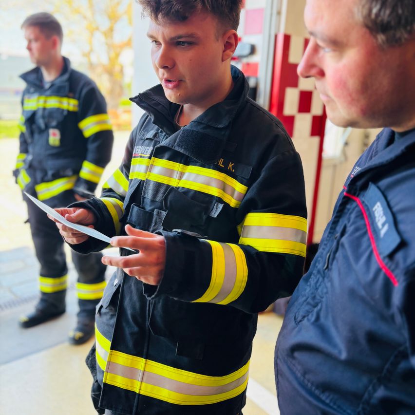 Drei Feuerwehrleute in Uniform, einer hält ein Papier, zwei unterhalten sich. Die zentrale Figur schaut auf das Papier und spricht. Im Hintergrund steht ein Feuerwehrauto.