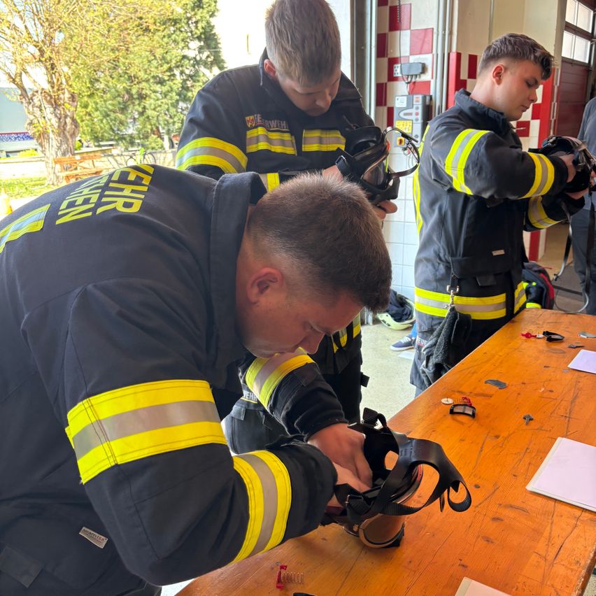 Drei Feuerwehrleute bereiten sich auf einen Notfall vor. Einer justiert einen Helm, während die anderen beobachten. Sie stehen in einem Raum mit einem Holztisch, Papieren und einem Baum draußen.
