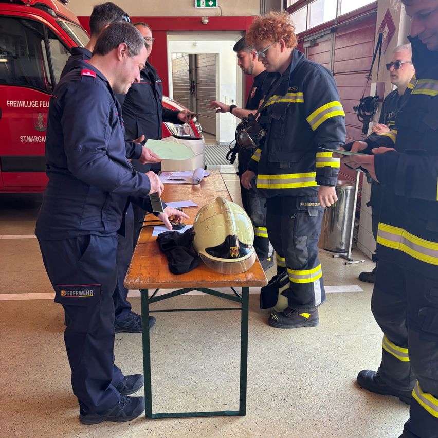 Eine Gruppe von Feuerwehrleuten steht um einen Tisch herum. Ein Mann hält ein Telefon und Papier, während andere zuschauen. Ein Feuerwehrhelm liegt auf dem Tisch. Hinter ihnen steht ein roter Feuerwehrwagen.