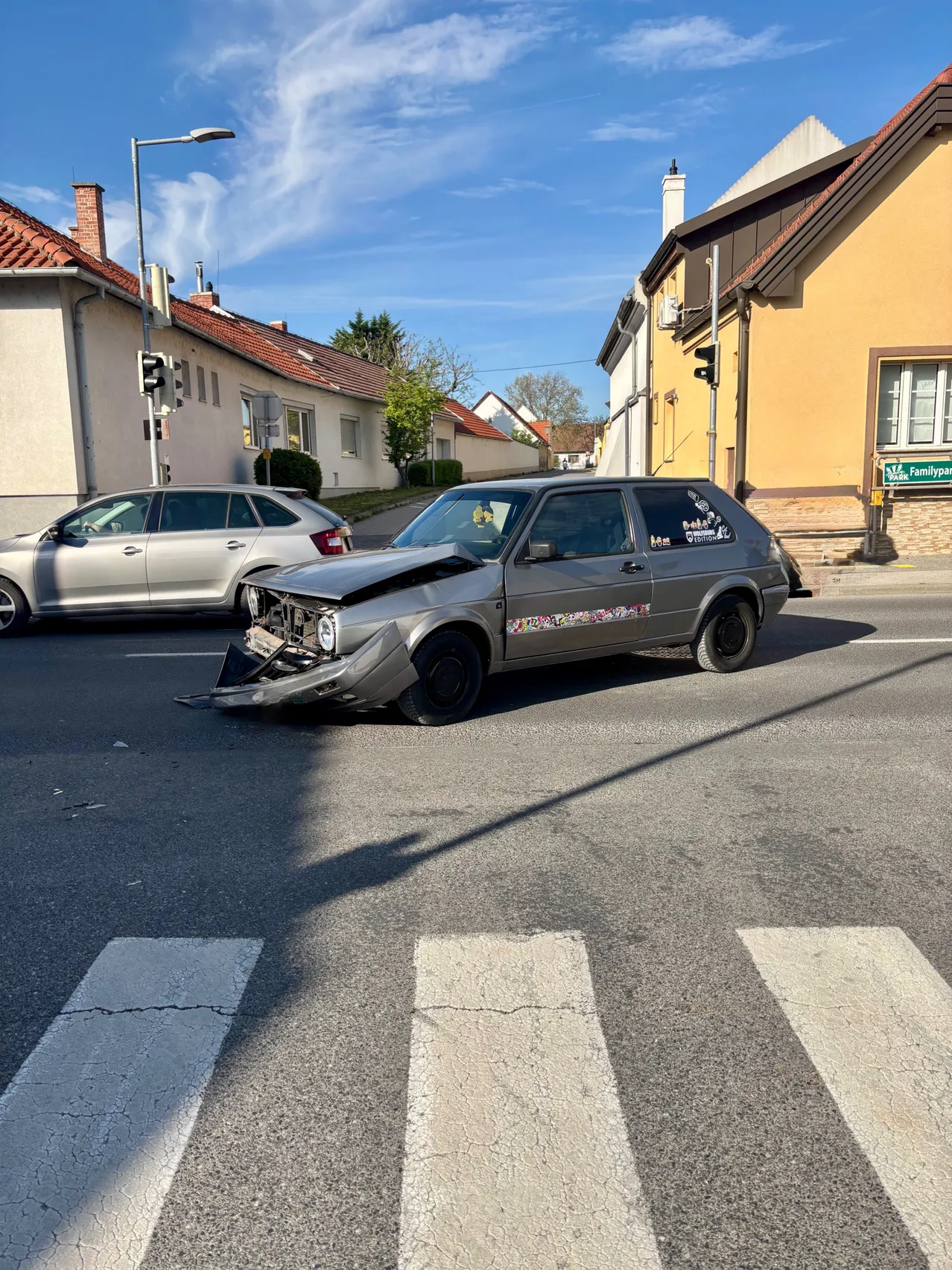 Ein graues Auto mit demolierter Front steht auf einer Straße. Ein silbernes Auto befindet sich dahinter. Gebäude säumen die Straße.