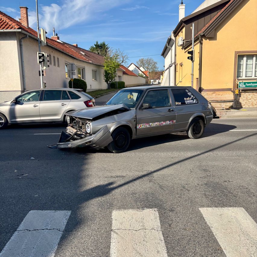 Ein graues Auto mit demolierter Front steht auf einer Straße. Ein silbernes Auto befindet sich dahinter. Gebäude säumen die Straße.
