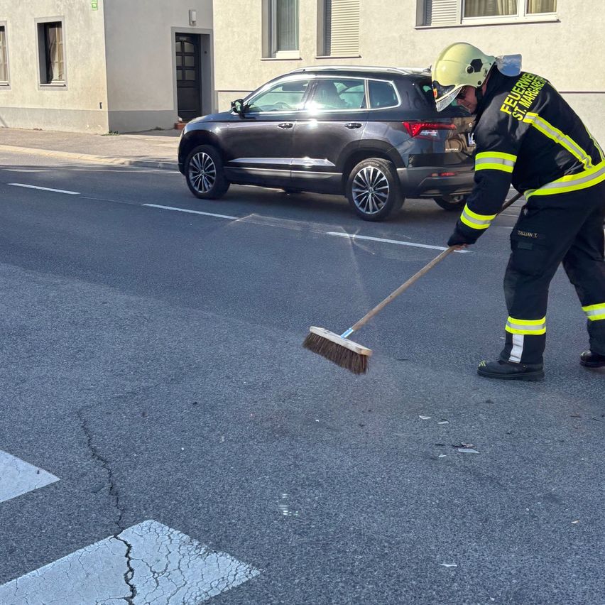 Ein Feuerwehrmann mit Reflexweste und Helm fegt die Straße mit einem Besen in der Nähe eines geparkten Autos vor einem Gebäude.