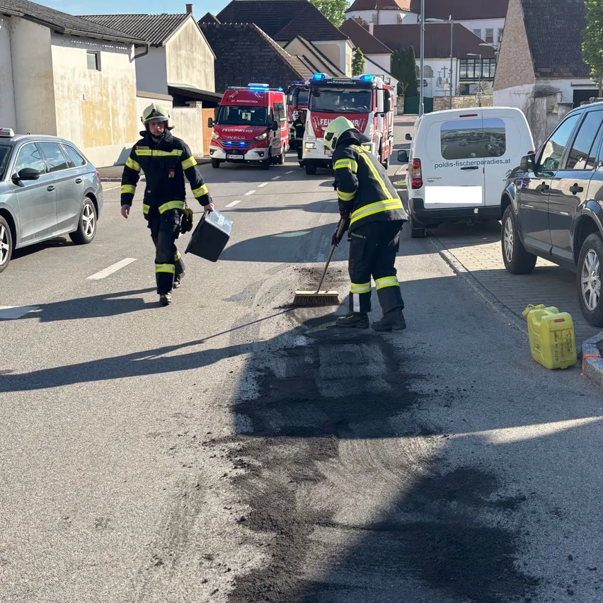 Zwei Feuerwehrleute reinigen eine Straße mit geparkten Feuerwehrwagen und Fahrzeugen in der Nähe. Ein Gebäude befindet sich links, und ein Lieferwagen und ein SUV befinden sich rechts.