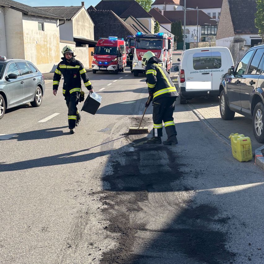 Zwei Feuerwehrleute reinigen eine Straße mit geparkten Feuerwehrwagen und Fahrzeugen in der Nähe. Ein Gebäude befindet sich links, und ein Lieferwagen und ein SUV befinden sich rechts.