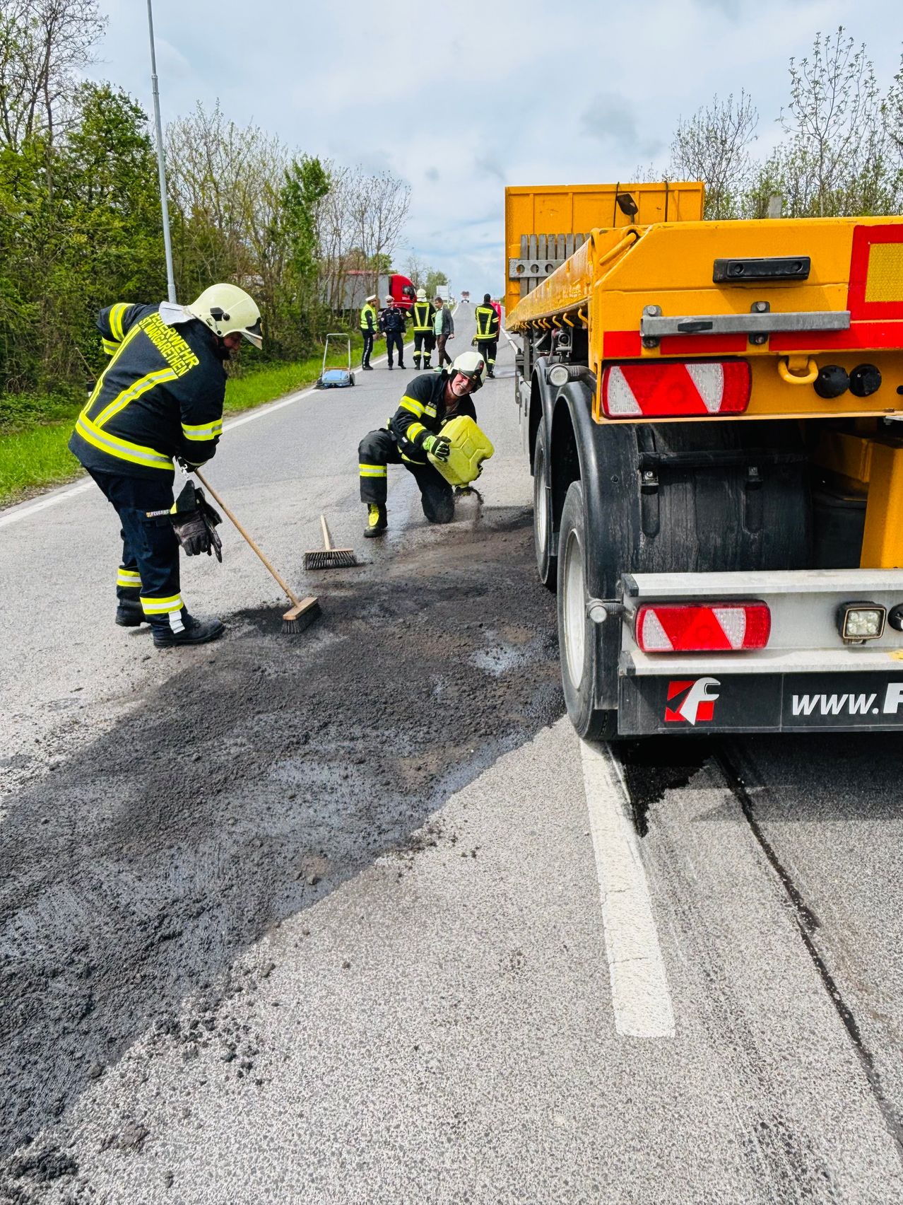 Feuerwehrleute reinigen eine Verschüttung auf der Straße. Einer kniet mit einem gelben Behälter, während der andere fegt. Ein gelber LKW ist in der Nähe geparkt.