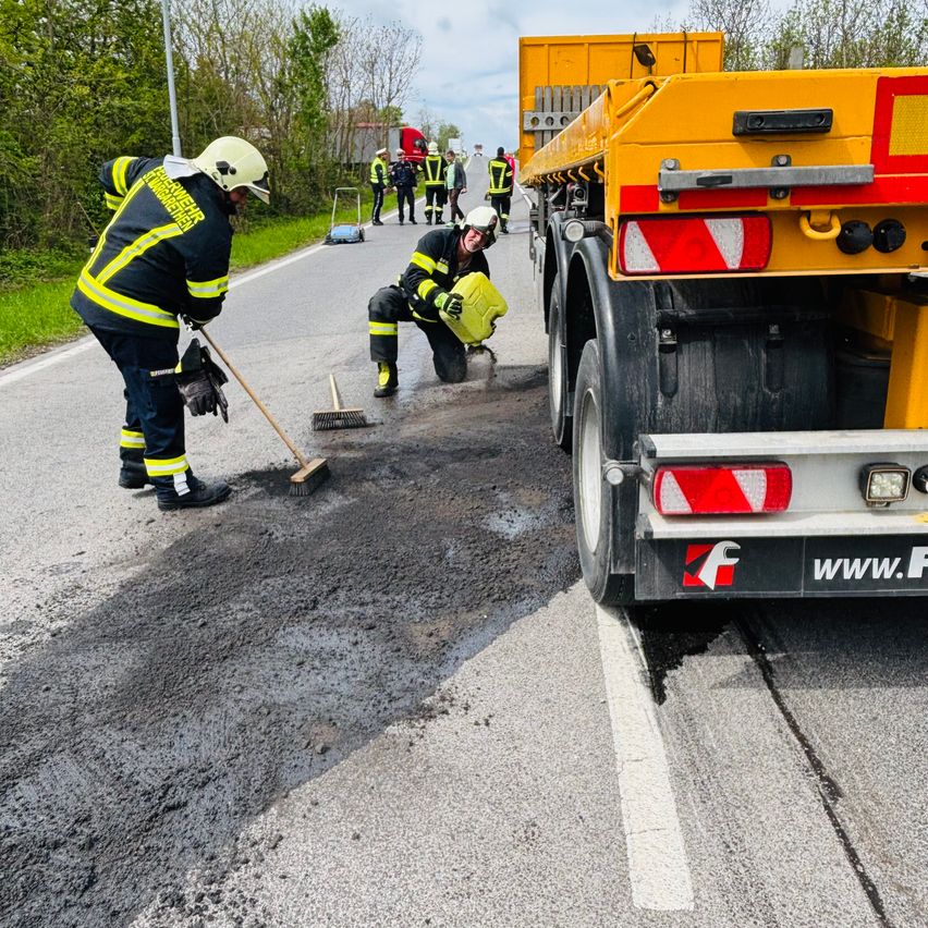 Feuerwehrleute reinigen eine Verschüttung auf der Straße. Einer kniet mit einem gelben Behälter, während der andere fegt. Ein gelber LKW ist in der Nähe geparkt.