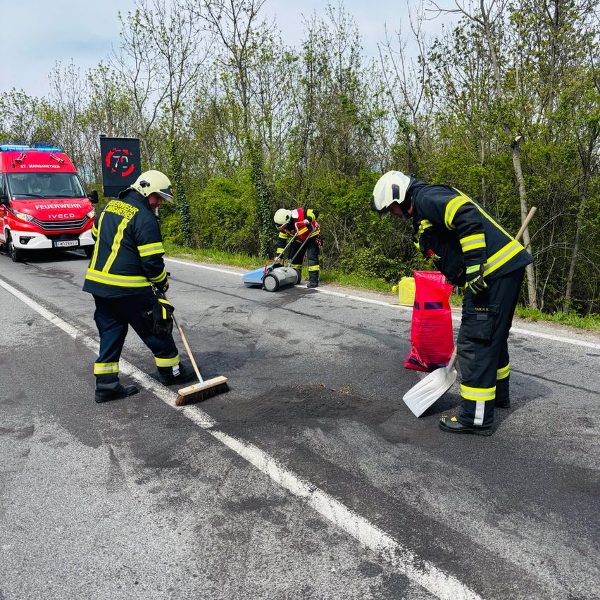 Feuerwehrleute reinigen die Straße nach einem Fahrzeugunfall, einer mit einem Besen und der andere mit einer Schaufel. Ein Krankenwagen steht hinter ihnen.