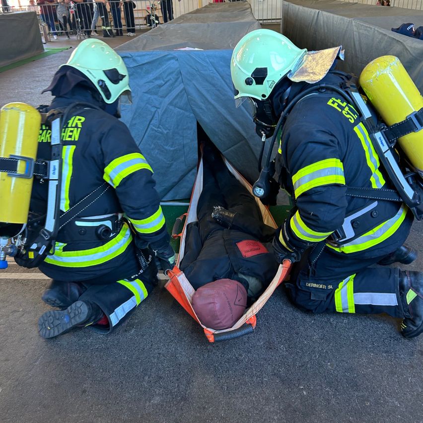 Zwei Feuerwehrleute üben Rettungsaktionen. Einer trägt eine Person auf einer Trage, während der andere sie stützt. Sie tragen Helme und tragen Sauerstoffflaschen.