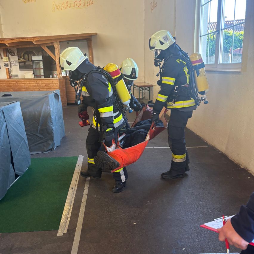Drei Feuerwehrleute tragen eine Puppe in einer orangefarbenen Tragehalle. Sie tragen Schutzausrüstung und Sauerstoffflaschen. Eine Person hält ein Klemmbrett, und ein Fenster zeigt eine Aussicht nach draußen.