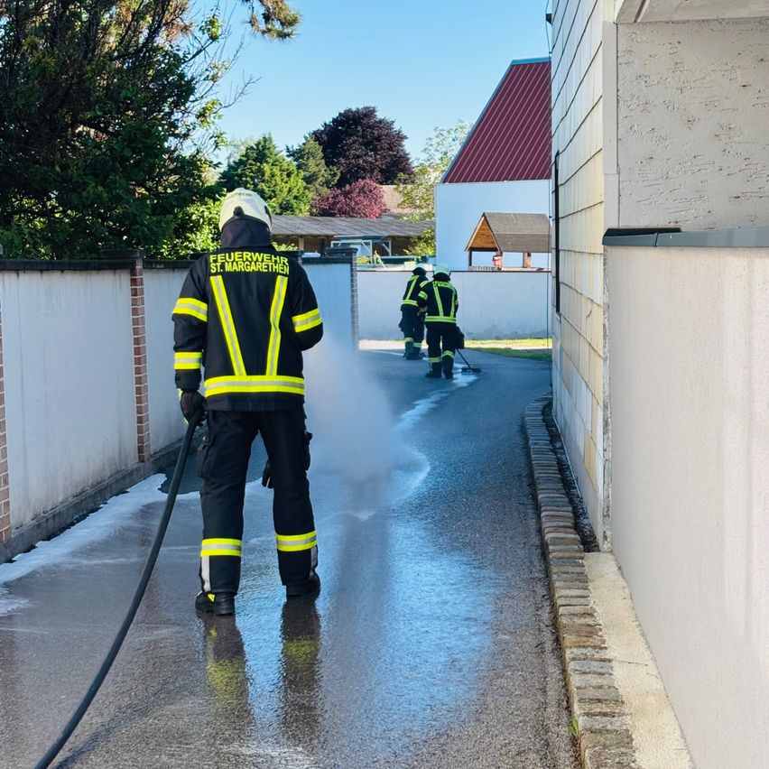 Feuerwehrleute in gelben und schwarzen Uniformen reinigen den Bürgersteig mit einem Schlauch. Ein Gebäude mit rotem Dach und ein Baum sind im Hintergrund.