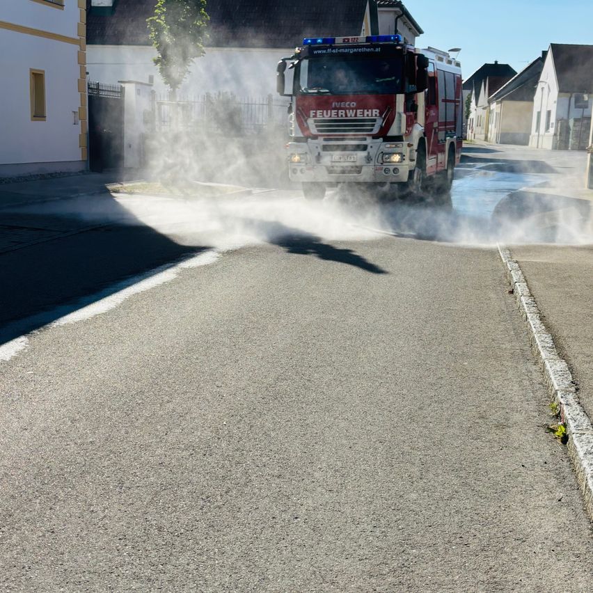 Ein Feuerwehrauto sprüht Wasser auf die Straße und erzeugt eine große Dampfwolke. Das Auto ist auf der Straße geparkt, mit Häusern auf beiden Seiten.