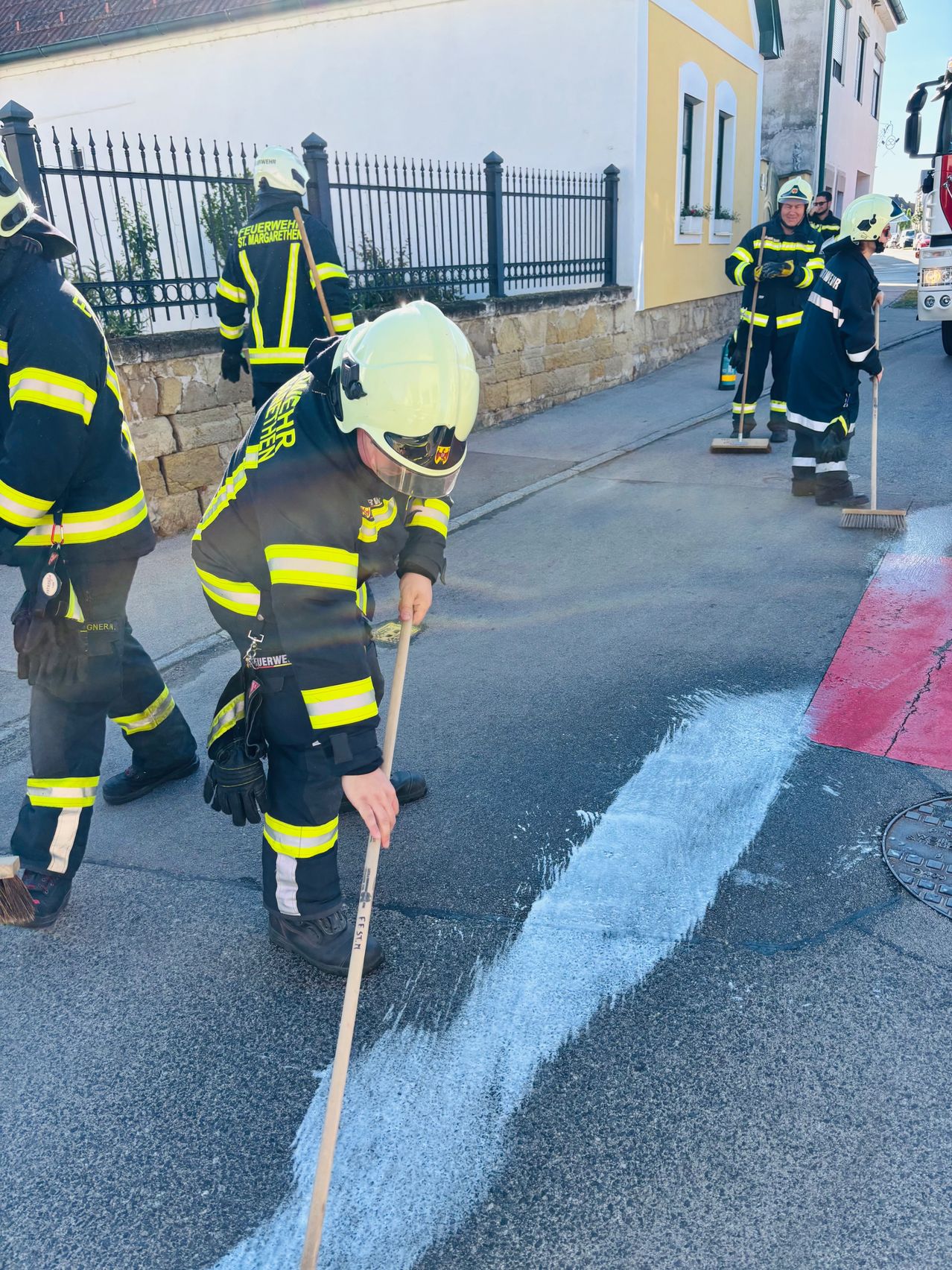 Feuerwehrleute in Uniform malen die Straße mit weißen und roten Markierungen. Eine Person benutzt einen Besen, um weißen Farbe zu verteilen.