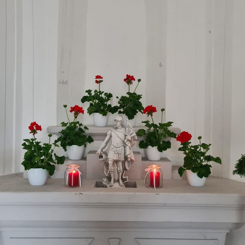 Ein weißer Regal mit einer Statue eines Soldaten zwischen zwei brennenden Kerzen, umgeben von weißen Töpfen mit roten Blumen.