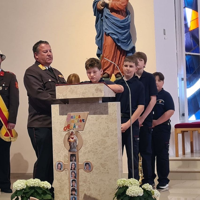 Ein Erwachsener in Uniform steht an einem Podest in einer Kirche, mit einer Statue dahinter. Mehrere Jungen in Uniform stehen in der Nähe.
