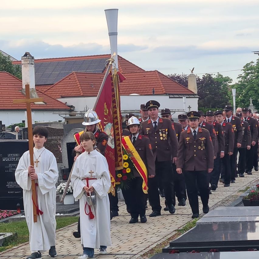 Eine feierliche Prozession von Militärangehörigen in Uniformen geht auf einem Friedhof, begleitet von zwei jungen Jungen, die Kreuze halten. Dahinter werden eine Flagge und eine Girlande getragen.