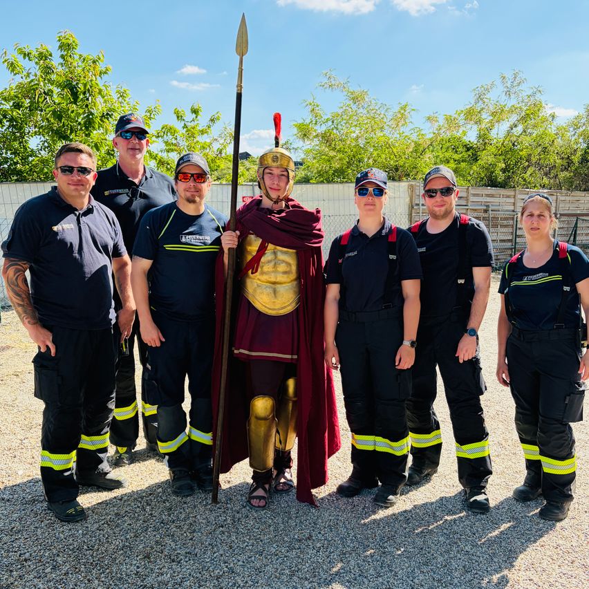 Eine Gruppe von Feuerwehrleuten in Uniform posiert für ein Foto mit einer Person, die als römischer Soldat verkleidet ist. Sie stehen auf einer Kiesfläche mit Bäumen und einem Zaun im Hintergrund.