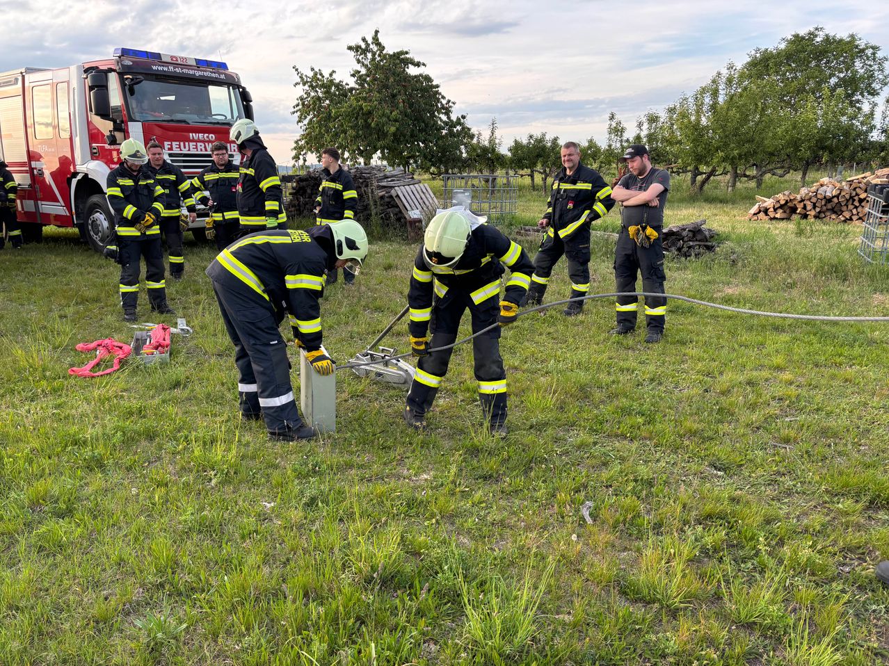 Eine Gruppe Feuerwehrleute in voller Ausrüstung führt eine Übung auf einer Grasfläche durch, wobei einige Werkzeuge halten. Dahinter steht ein Feuerwehrwagen und ein Feuerwehrmann.
