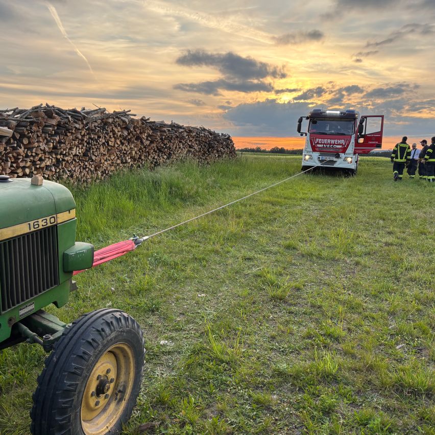 Ein Feuerwehrauto mit offener Tür ist mit einem Traktor mit der Nummer 1630 auf einer Wiese mit Holzstapel verbunden.