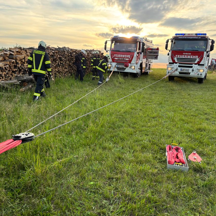 Feuerwehrleute bereiten sich auf das Ziehen mit Feuerwehrfahrzeugen auf einer Wiese vor, mit einem Stapel Holzscheite im Hintergrund.