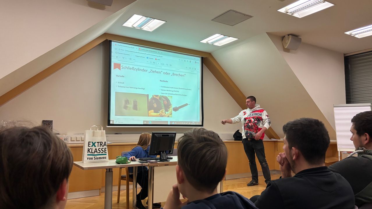 Ein Mann steht vor einer Projektionsleinwand und präsentiert eine Folie mit Text. Er trägt eine Jacke mit rotem und weißem Design. Mehrere Schüler sitzen vor ihm an Tischen.