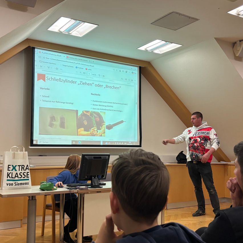 Ein Mann steht vor einer Projektionsleinwand und präsentiert eine Folie mit Text. Er trägt eine Jacke mit rotem und weißem Design. Mehrere Schüler sitzen vor ihm an Tischen.