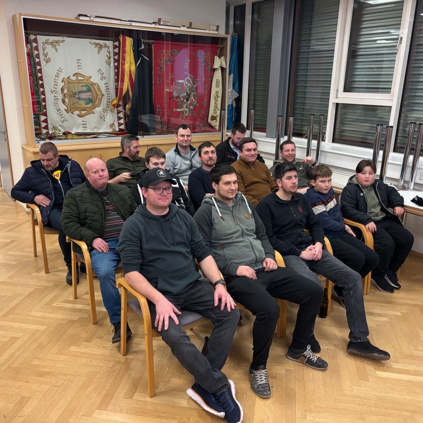 Eine Gruppe von Männern und Jungen sitzt auf Stühlen in einem Raum, einige tragen Jacken und Mützen, mit einem Banner im Hintergrund.