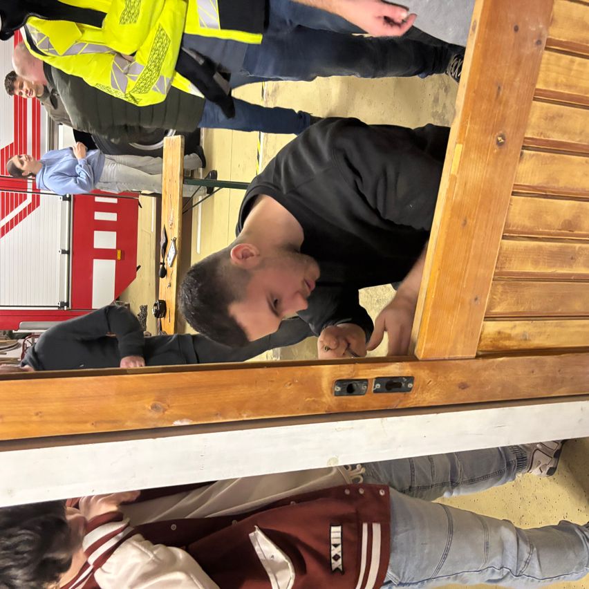 Ein Mann in einem schwarzen Shirt lehnt sich an eine Holztür. Hinter ihm steht eine weitere Person mit einer gelben Weste und Hose. Im Hintergrund ist ein Mann in einem blauen Hemd und Hose zu sehen. Der Boden ist aus Holz.