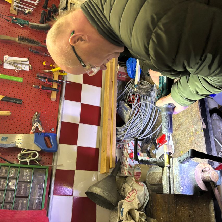 Ein Mann mit Brille und grüner Jacke arbeitet in einer Werkstatt an einer Maschine. Die Werkstatt hat einen rot-weiß karierten Boden.