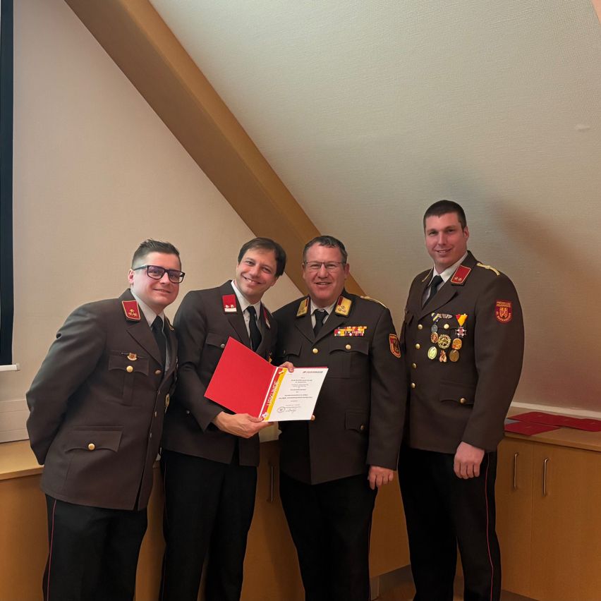 Vier Männer in Militäruniformen stehen zusammen und lächeln. Der Mann in der Mitte hält eine rote Mappe und ein Zertifikat. Die anderen tragen Medaillen. Hinter ihnen befindet sich eine weiße Wand mit einem Fenster und einem Holzschrank.