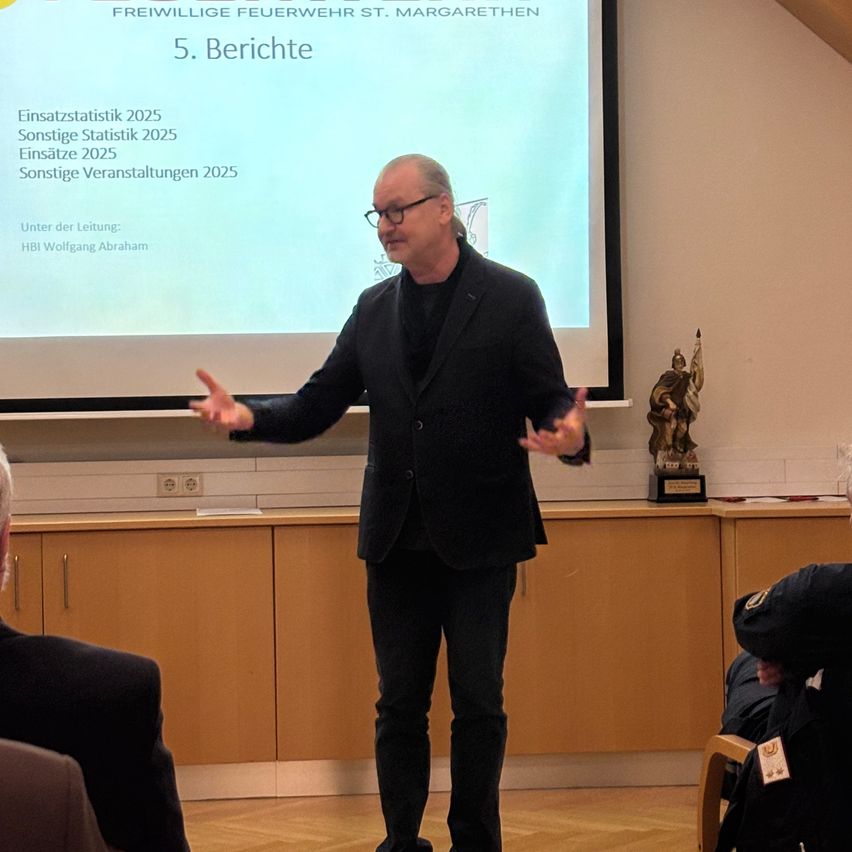 Ein Mann hält eine Präsentation vor einer Projektionsleinwand. Er steht in einem Klassenzimmer, und ein Pokal ist rechts.