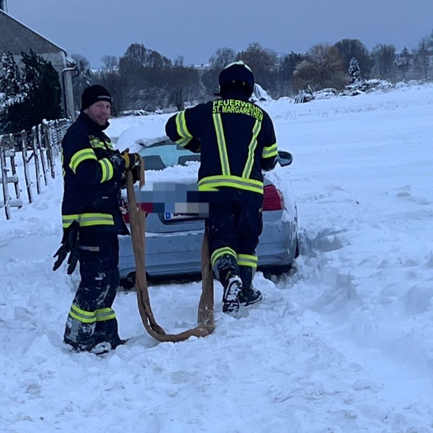 Zwei Feuerwehrleute versuchen, ein im Schnee steckendes Auto zu retten, einer hält eine Seil. Das Auto ist teilweise vergraben.