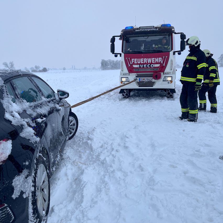 Ein Auto wird von einem Feuerwehrauto auf einem verschneiten Feld gezogen. Zwei Feuerwehrleute beobachten es.