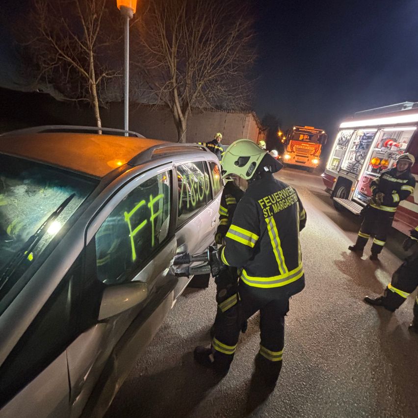 Feuerwehrleute arbeiten an einem beschädigten Auto mit gelben Markierungen und dem Wort 'FF' auf der Scheibe. Ein weiterer Feuerwehrmann steht neben einem Feuerwehrwagen.