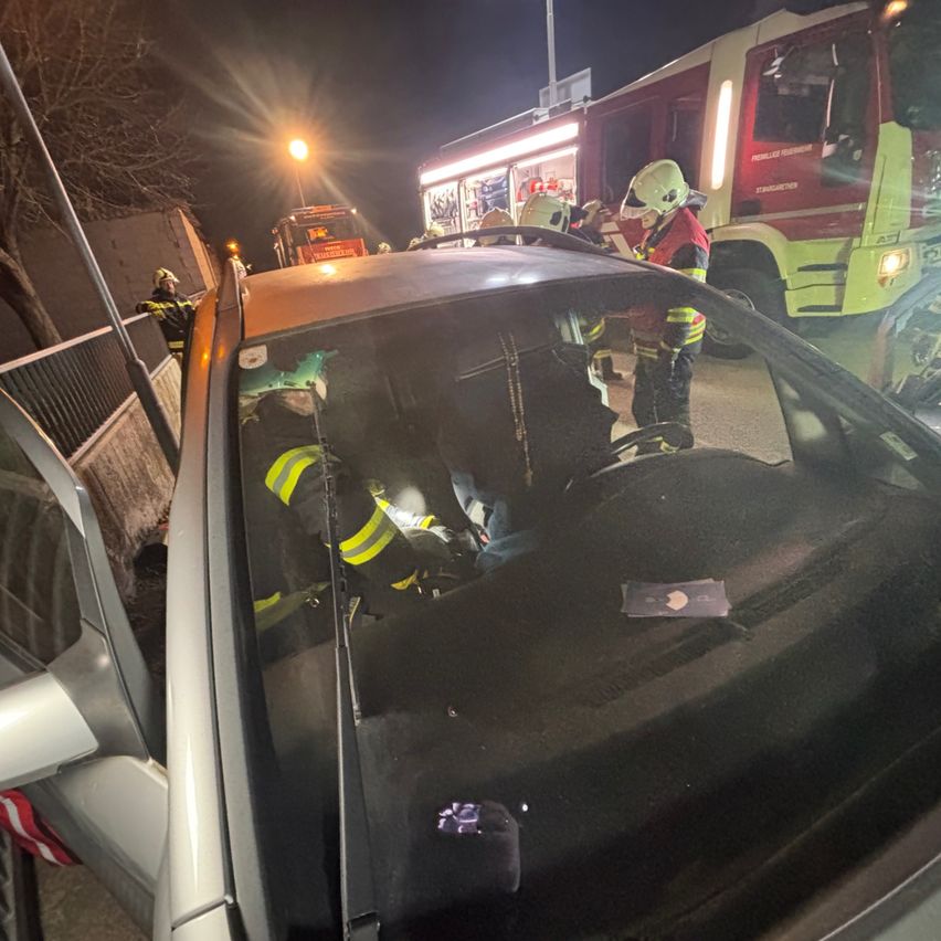 Feuerwehrleute in Schutzkleidung arbeiten nachts an einem beschädigten Fahrzeug. Ein Feuerwehrauto steht dahinter.