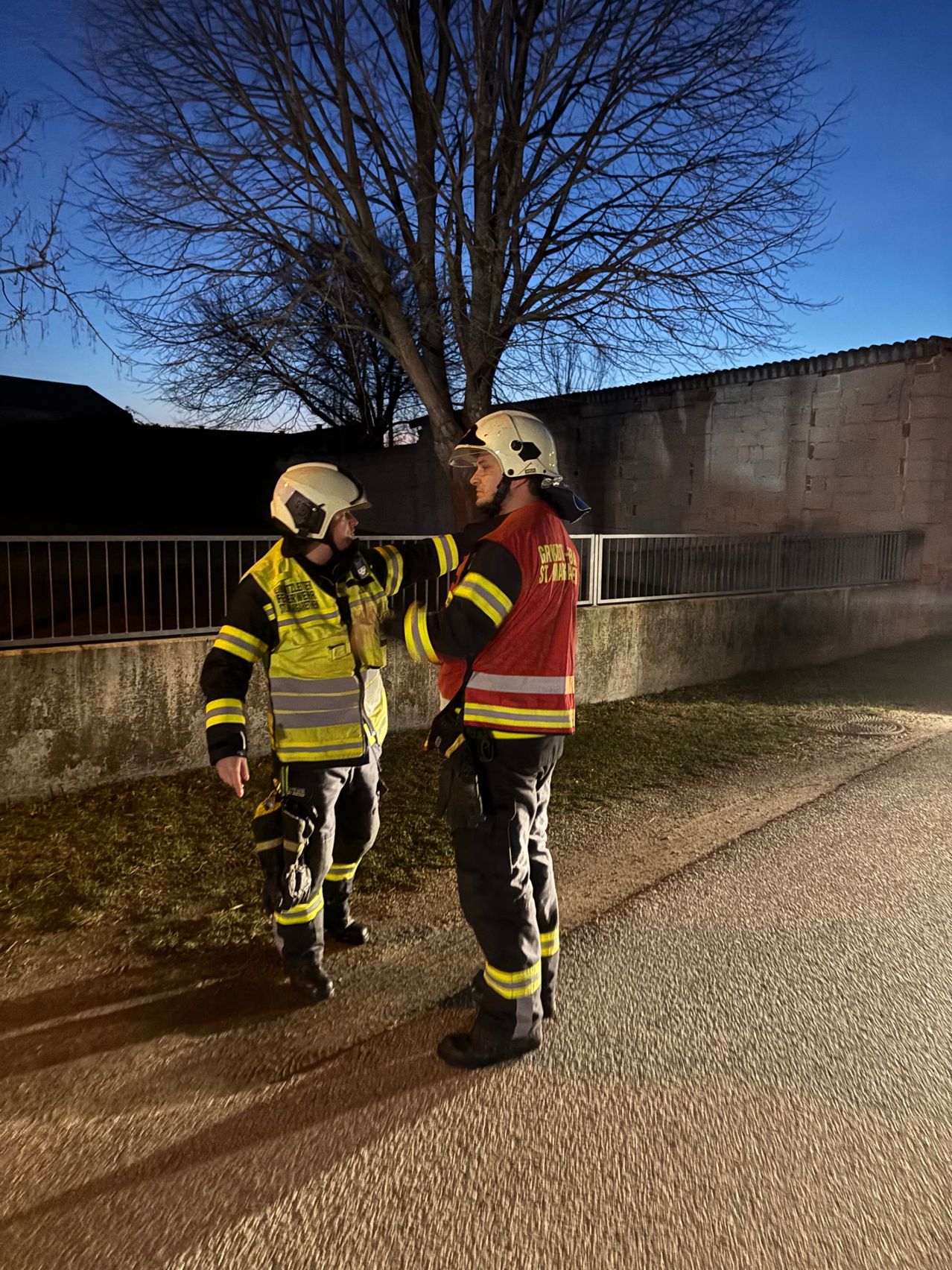 Zwei Feuerwehrleute stehen auf einer Straße, einer in einer gelben Jacke und der andere in einer roten Jacke, beide tragen Helme. Sie stehen neben einer Mauer mit einem Baum dahinter.
