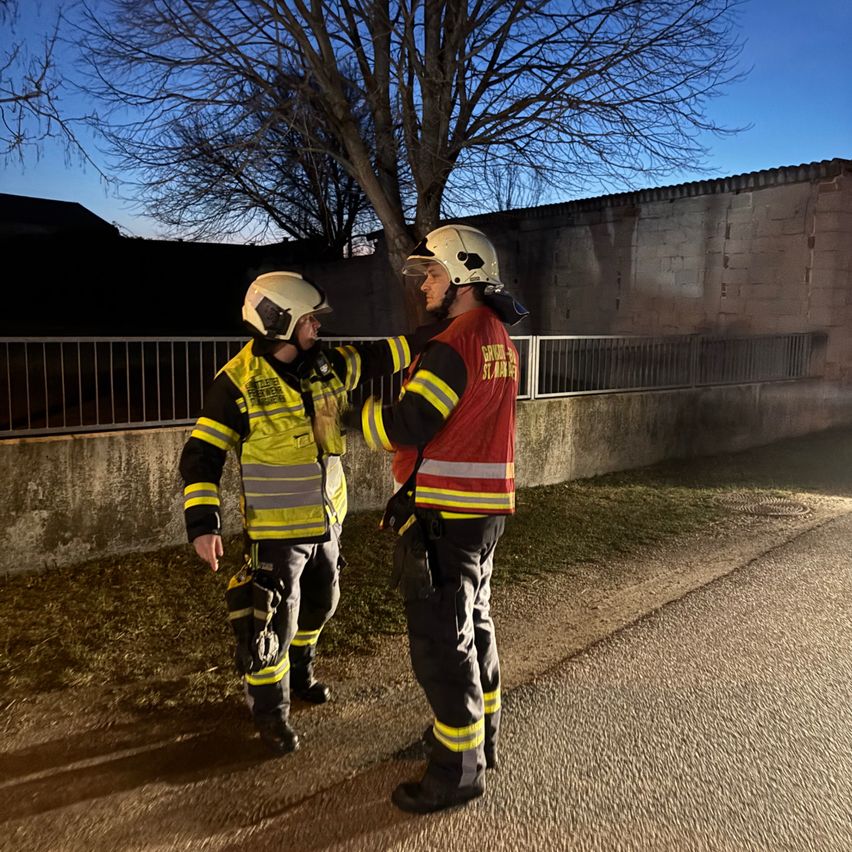 Zwei Feuerwehrleute stehen auf einer Straße, einer in einer gelben Jacke und der andere in einer roten Jacke, beide tragen Helme. Sie stehen neben einer Mauer mit einem Baum dahinter.
