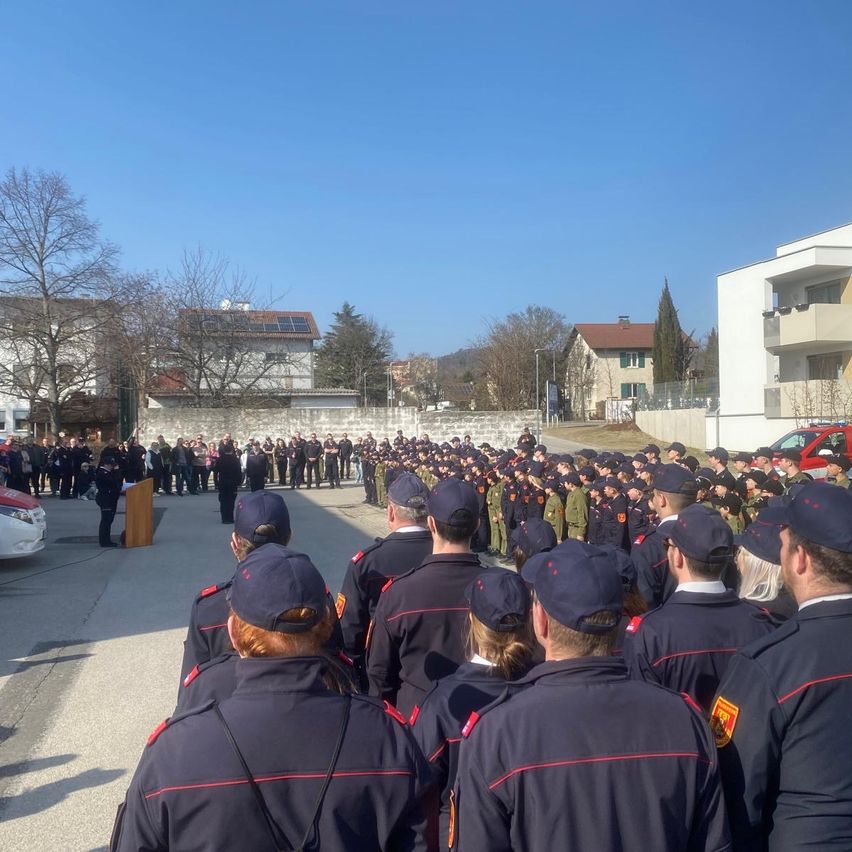 Eine große Versammlung von Menschen in Uniform trifft sich auf einem Freigelände, mit einem Podium und einer sprechenden Person im Vordergrund. Im Hintergrund sind mehrere Gebäude mit Solarpanelen und Bäumen zu sehen.