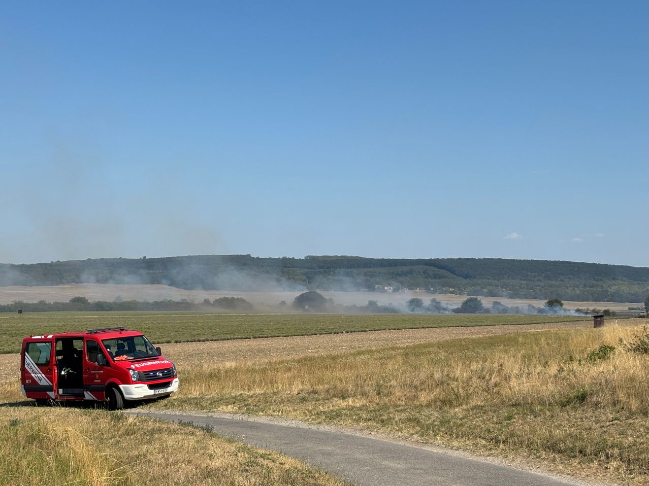 Ein roter Feuerwehrwagen steht am Straßenrand, mit einem Feld und entfernten Hügeln im Hintergrund, und Rauch steigt von den Hügeln auf.