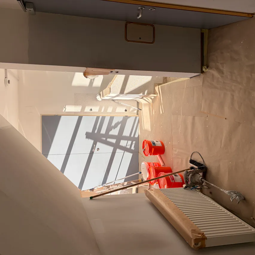 Ein Baustelle mit einer weißen Wand und einer Tür. Zwei orangefarbene Eimer stehen auf dem Boden. Ein weißer Heizkörper befindet sich auf der rechten Seite.
